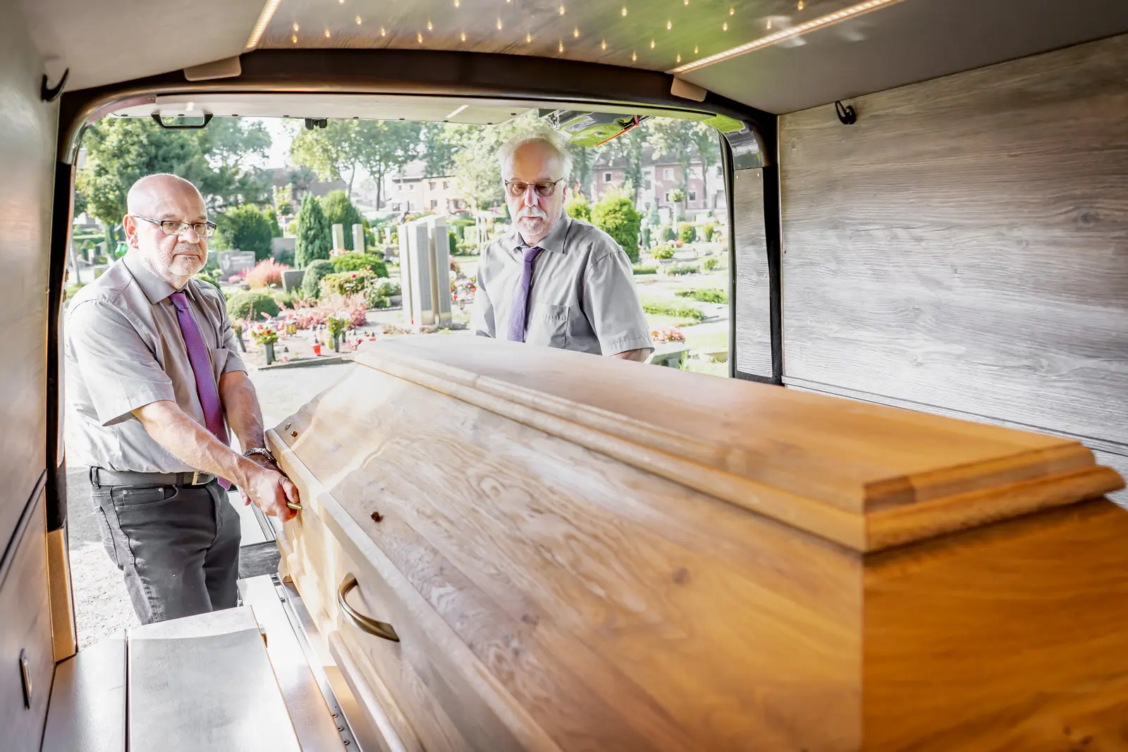 Bestattungshaus Ludger Schlüter | Zwei Mitarbeiter überführen mit dem firmeneigenen Leichenwagen einen Sarg zum Friedhof.